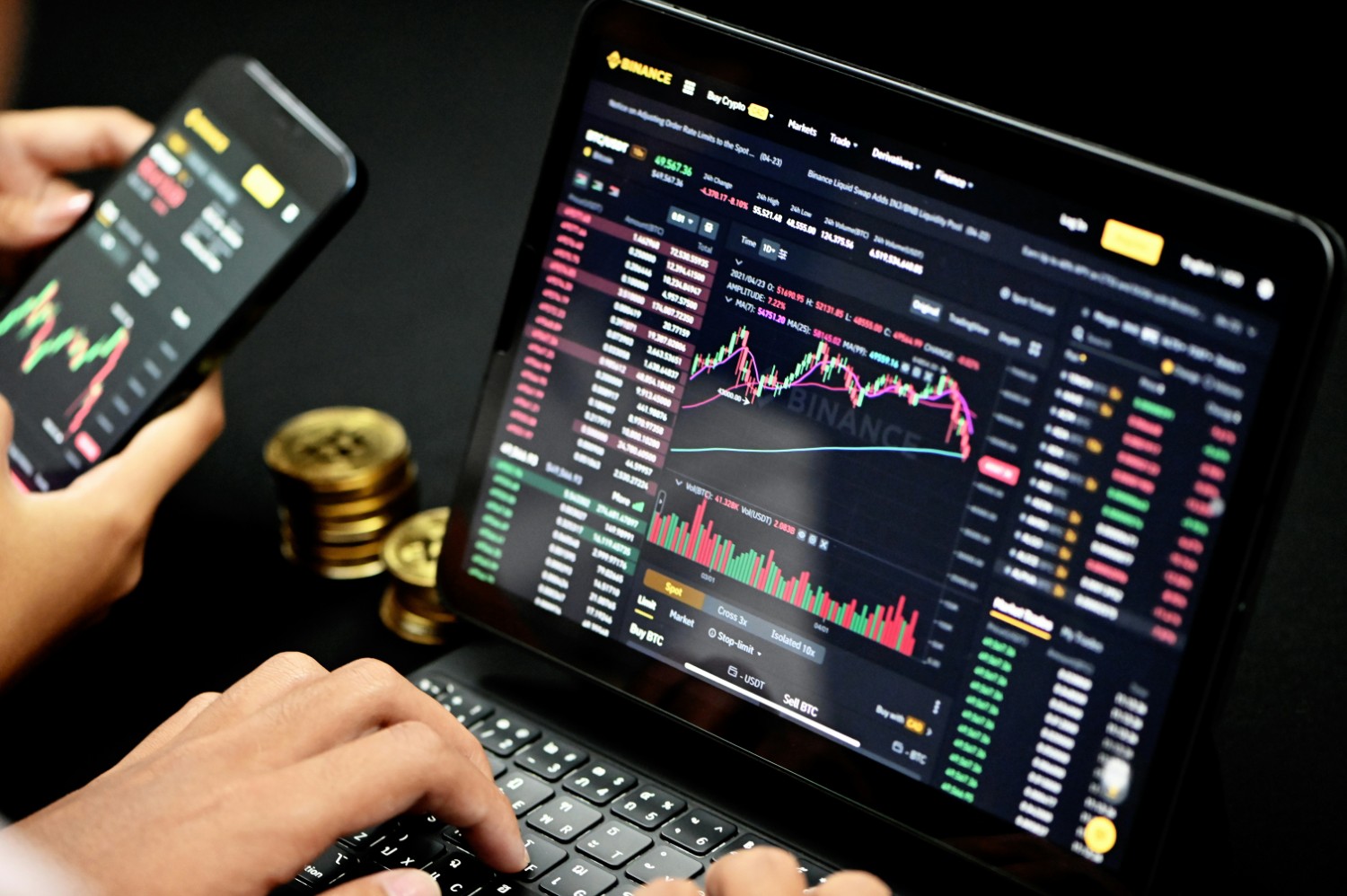 A trader analyzing cryptocurrency market trends on Binance using a laptop and smartphone, with stacks of Bitcoin coins in the background.